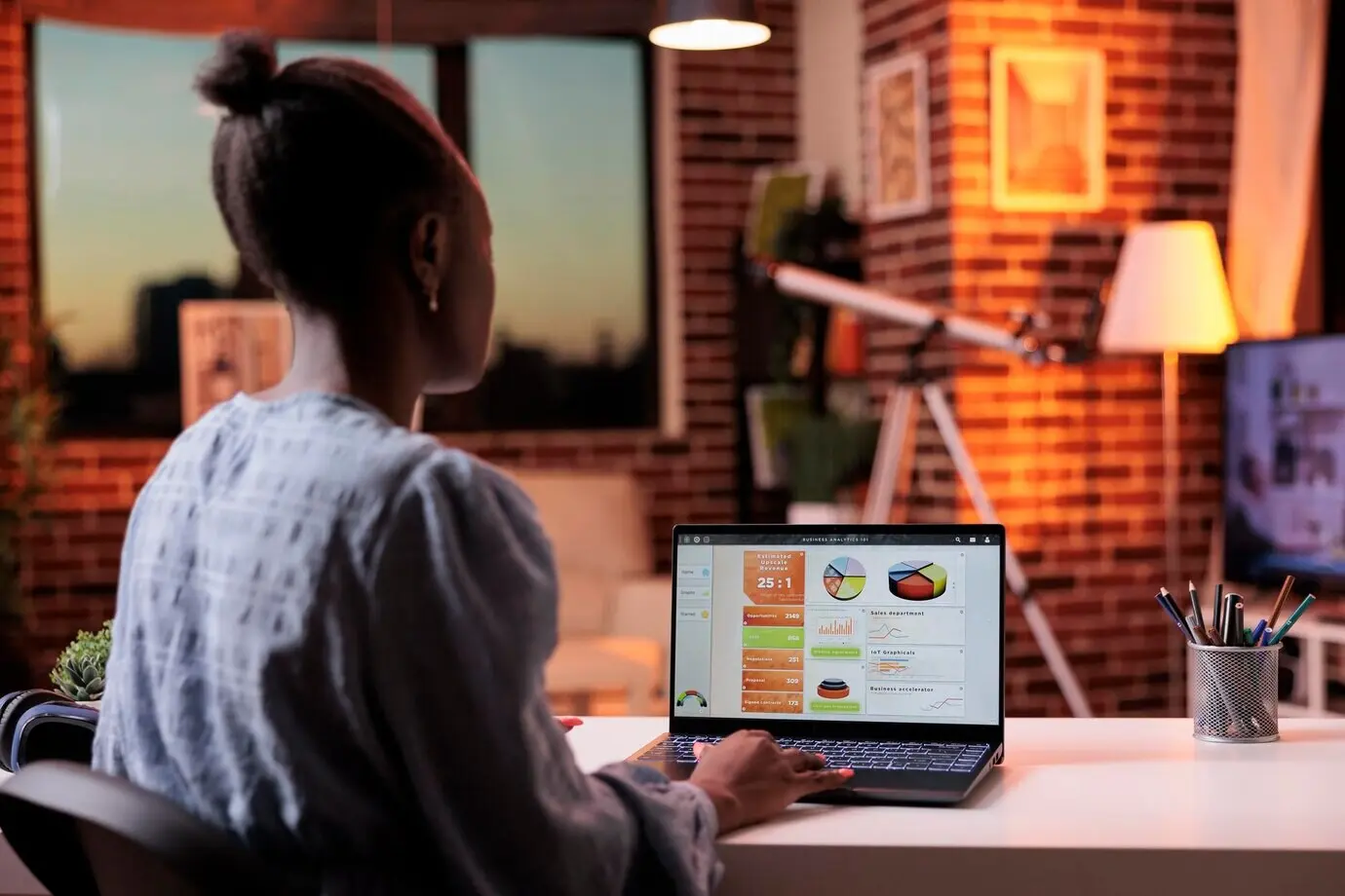 Geschäftsfrau in Rückansicht betrachtet auf einem Laptopbildschirm Statistiken aus der Marketingforschung des Unternehmens. Remote tätige Analystin analysiert im Homeoffice am Abend Diagramme und Kreisdiagramme am Computer.